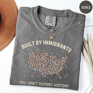 May include: A gray Comfort Colors t-shirt with the text "BUILT BY IMMIGRANTS" and "YOU CAN'T DEPORT HISTORY". The shirt features a graphic of the United States map filled with diverse figures. A silver necklace with a pendant is draped across the shirt.