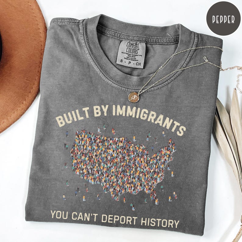 May include: A gray Comfort Colors t-shirt with the text "BUILT BY IMMIGRANTS" and "YOU CAN'T DEPORT HISTORY". The shirt features a graphic of the United States map filled with diverse figures. A silver necklace with a pendant is draped across the shirt.