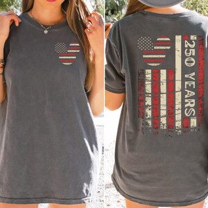 May include: Charcoal grey t-shirt featuring a distressed American flag design. The front has a Mickey Mouse head with stars and stripes. The back shows a larger flag design with "250 YEARS" text. A casual, patriotic tee.