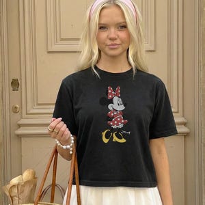 May include: Black t-shirt featuring a Minnie Mouse graphic in red, white, and yellow. The shirt is paired with a white skirt and a pink headband. A brown tote bag with bread is in the foreground.