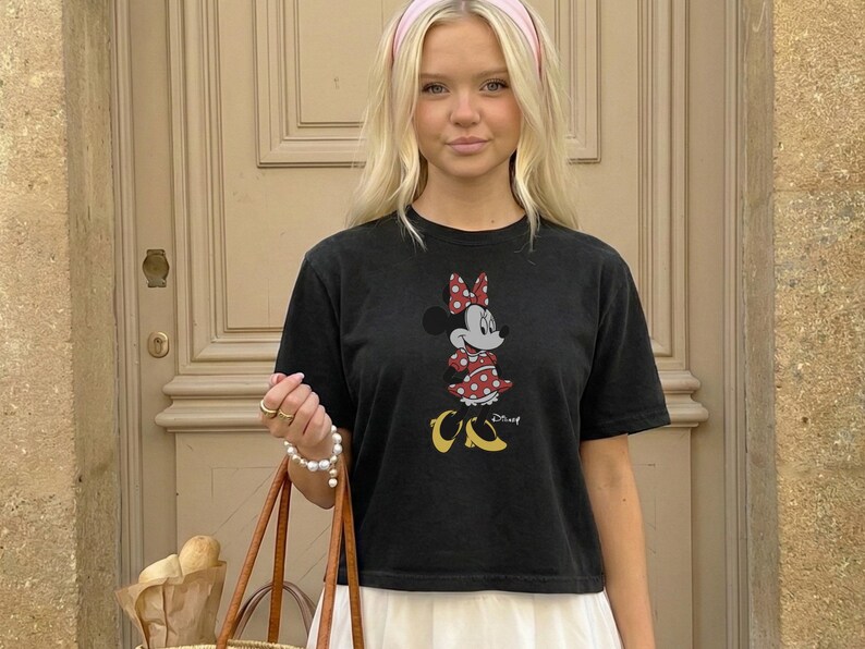 May include: Black t-shirt featuring a Minnie Mouse graphic in red, white, and yellow. The shirt is paired with a white skirt and a pink headband. A brown tote bag with bread is in the foreground.