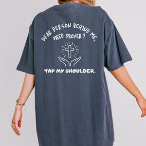 May include: A dark blue t-shirt with white text that reads "Dear person behind me, need prayer? Tap my shoulder."  The text is centered on the shirt and includes a graphic of a cross with hands reaching up towards it.