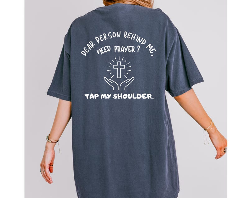 May include: A dark blue t-shirt with white text that reads "Dear person behind me, need prayer? Tap my shoulder."  The text is centered on the shirt and includes a graphic of a cross with hands reaching up towards it.