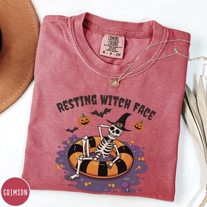 May include: Crimson-colored t-shirt with the phrase "RESTING WITCH FACE" and a Halloween-themed graphic. The graphic features a skeleton wearing a witch hat, lounging in an orange and black inner tube, surrounded by pumpkins, bats, and candy. A silver necklace is draped across the shirt.