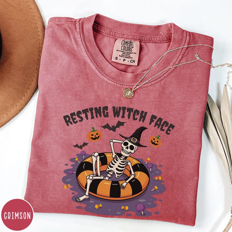 May include: Crimson-colored t-shirt with the phrase "RESTING WITCH FACE" and a Halloween-themed graphic. The graphic features a skeleton wearing a witch hat, lounging in an orange and black inner tube, surrounded by pumpkins, bats, and candy. A silver necklace is draped across the shirt.