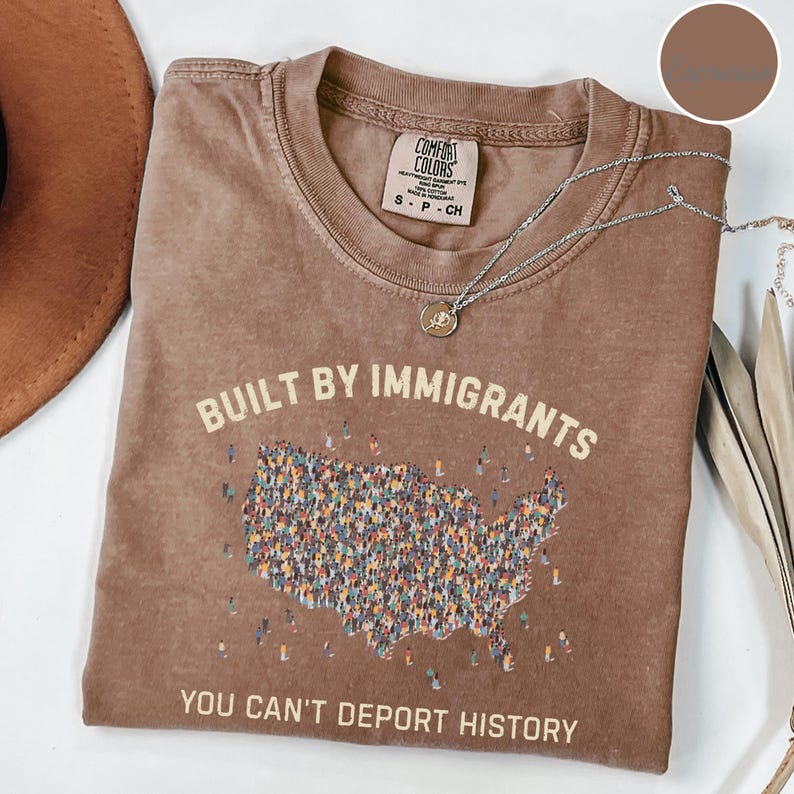 May include: Brown t-shirt with the text "BUILT BY IMMIGRANTS" and "YOU CAN'T DEPORT HISTORY". The graphic on the shirt is a map of the United States filled with small figures. A silver necklace with a pendant is on the shirt.