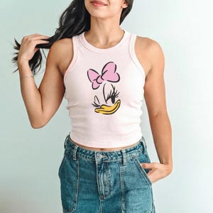 Op de afbeelding: Een lichtroze tanktop met een cartoonafbeelding van een roze strik, wimpers en een gele snavel. De tanktop is geribbeld en wordt gecombineerd met een blauwe spijkerbroek.
