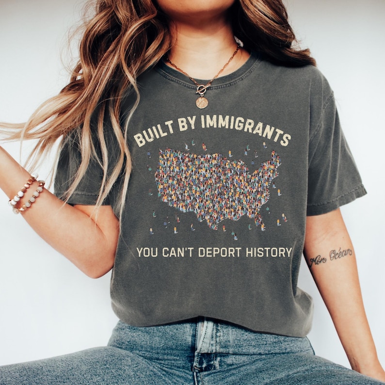 May include: Dark gray t-shirt with the text "BUILT BY IMMIGRANTS" above a map of the United States filled with tiny figures. Below the map, the text reads "YOU CAN'T DEPORT HISTORY".