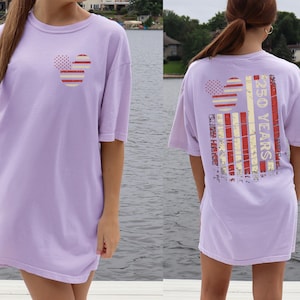 May include: A lilac t-shirt with a Mickey Mouse-style design on the front and back. The design incorporates red and white stripes and dots. The back of the shirt displays the text "250 YEARS" in a vertical arrangement.