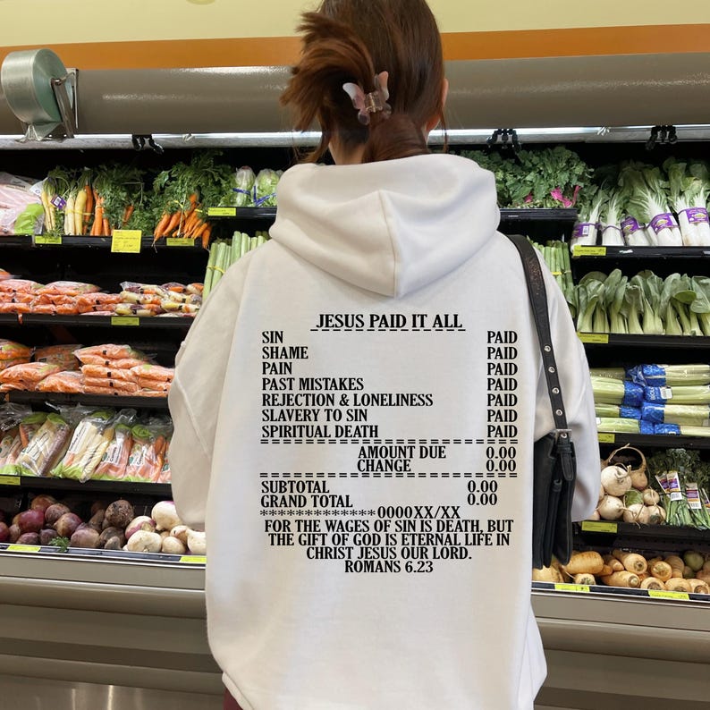 May include: White hooded sweatshirt with a black graphic print that reads "JESUS PAID IT ALL" and lists various sins with the word "PAID" next to each. The graphic also includes a receipt-like section with the words "AMOUNT DUE", "CHANGE", "SUBTOTAL", and "GRAND TOTAL" with the amount "0.00" listed next to each. The text "FOR THE WAGES OF SIN IS DEATH, BUT THE GIFT OF GOD IS ETERNAL LIFE IN CHRIST JESUS OUR LORD. ROMANS 6.23" is printed below the receipt.