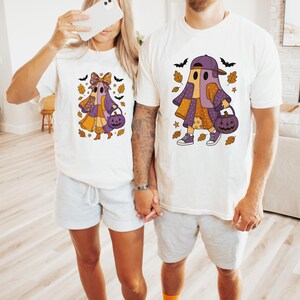 Könnte beinhalten: Weiße T-Shirts mit einem Halloween-Geister-Design. Der Geist ist aus Patchwork in Orange, Lila und Gelb, mit einer Kürbistasche. Das Design umfasst Fledermäuse und Herbstblätter.