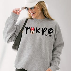 May include: Gray sweatshirt with the word "TOKYO" in black script, featuring a Minnie Mouse graphic and the year "2026". The sweatshirt is a crew neck style.