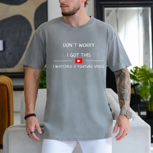 May include: Gray t-shirt with the text "DON'T WORRY I GOT THIS" above a white line and a YouTube play button, followed by "I WATCHED A YOUTUBE VIDEO". The shirt is short-sleeved and appears to be made of a soft material.