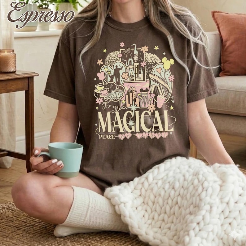 May include: A dark brown short-sleeve t-shirt featuring a multi-colored graphic print of a castle, fireworks, and various park icons. The text reads The Most MAGICAL PLACE ON EARTH. The design includes soft pink, yellow, and white accents.