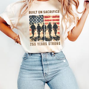 May include: Cream-colored t-shirt featuring a graphic of the American flag with soldiers and the text "BUILT ON SACRIFICE" and "250 YEARS STRONG". The design is printed on a soft, casual tee.