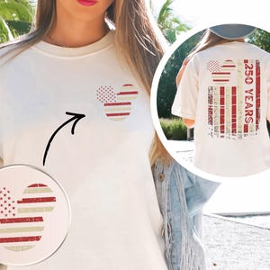 May include: Cream-coloured t-shirt featuring a patriotic Mickey Mouse design on the front and back. The design incorporates red and white stripes and stars, with the text "250 YEARS" on the back. The shirt has a relaxed fit.