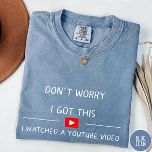 May include: A blue t-shirt with the text "DON'T WORRY I GOT THIS I WATCHED A YOUTUBE VIDEO". The shirt has a small red and white YouTube logo. A gold necklace with a pendant is on top of the shirt.