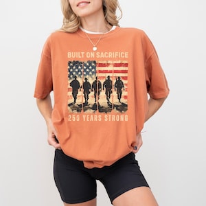 May include: Burnt orange t-shirt with a graphic of the American flag and silhouettes of soldiers. The text "BUILT ON SACRIFICE" and "250 YEARS STRONG" are printed above and below the flag design. The shirt is a relaxed fit.
