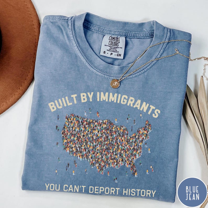 May include: A blue Comfort Colors t-shirt with the text "BUILT BY IMMIGRANTS" and "YOU CAN'T DEPORT HISTORY". The design includes a map of the United States filled with tiny figures. A gold necklace is draped across the shirt.