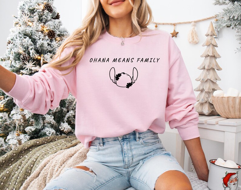 May include: A pale pink sweatshirt featuring the words "OHANA MEANS FAMILY" in black, with a black character graphic below. The long-sleeved top is made of a soft material and is suitable for casual wear.