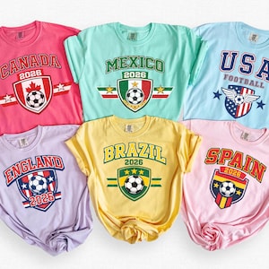 May include: Six short-sleeve t-shirts in pink, mint green, light blue, lavender, yellow, and light pink. Each shirt features a graphic with a country name, the year 2026, and a soccer ball emblem representing Canada, Mexico, USA, England, Brazil, and Spain.