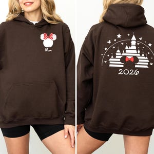 Puede incluir: Una sudadera con capucha marr&oacute;n con un bolsillo delantero. La parte delantera presenta un dise&ntilde;o de Minnie Mouse con un lazo rojo y la palabra "Mom". La parte trasera muestra un dise&ntilde;o de castillo con el a&ntilde;o "2026".