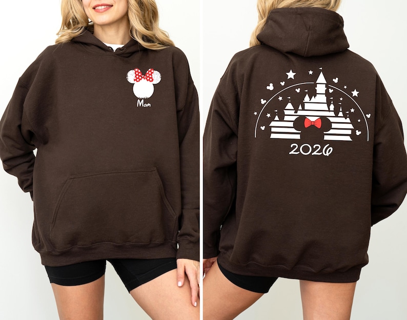 Puede incluir: Una sudadera con capucha marr&oacute;n con un bolsillo delantero. La parte delantera presenta un dise&ntilde;o de Minnie Mouse con un lazo rojo y la palabra "Mom". La parte trasera muestra un dise&ntilde;o de castillo con el a&ntilde;o "2026".