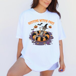 May include: White t-shirt with the text "RESTING WITCH FACE" in orange. The graphic features a skeleton wearing a witch hat, floating in a black and orange inner tube, with pumpkins and bats. Halloween-themed apparel.