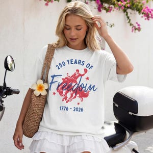 May include: White t-shirt with a red and blue graphic of a person riding a horse, with the text "250 Years of Freedom 1776-2026". The shirt is a casual style.
