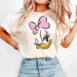 May include: Cream-colored t-shirt featuring a cartoon illustration of Daisy Duck. The design includes a pink bow, black eyelashes, and a yellow beak. The shirt is made of soft material and has rolled-up sleeves.