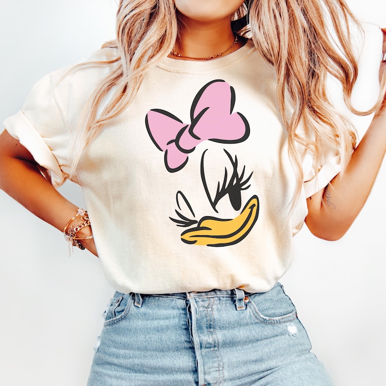 May include: Cream-colored t-shirt featuring a cartoon illustration of Daisy Duck. The design includes a pink bow, black eyelashes, and a yellow beak. The shirt is made of soft material and has rolled-up sleeves.