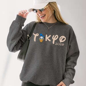 May include: Dark gray sweatshirt featuring the word "TOKYO" in a stylized font, with a cartoon Donald Duck graphic and the year "2026". The sweatshirt has long sleeves and a crew neck. The person in the image is wearing a white cap and sunglasses.