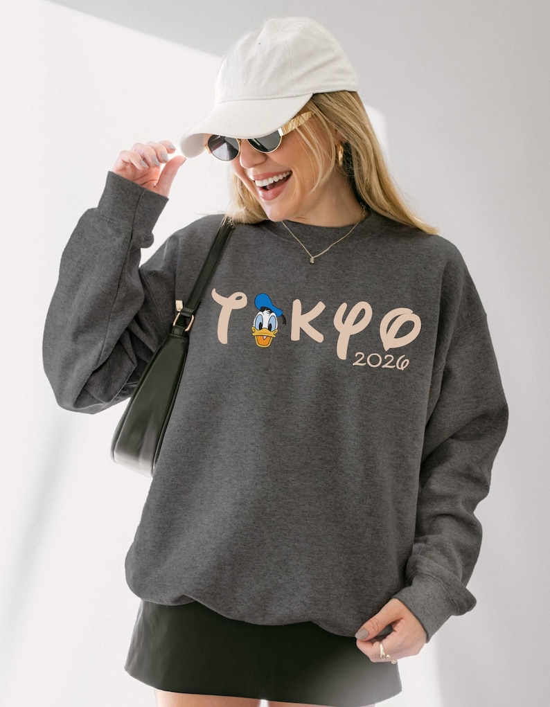 May include: Dark gray sweatshirt featuring the word "TOKYO" in a stylized font, with a cartoon Donald Duck graphic and the year "2026". The sweatshirt has long sleeves and a crew neck. The person in the image is wearing a white cap and sunglasses.
