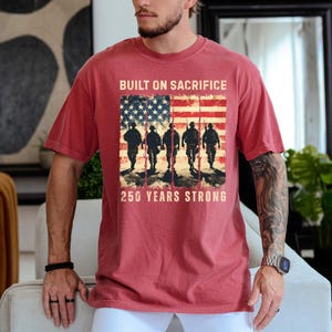 May include: Red t-shirt with a graphic featuring the American flag and silhouettes of soldiers. The text reads "BUILT ON SACRIFICE" above the flag and "250 YEARS STRONG" below. The shirt is a solid red color.