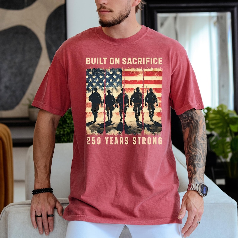 May include: Red t-shirt with a graphic featuring the American flag and silhouettes of soldiers. The text reads "BUILT ON SACRIFICE" above the flag and "250 YEARS STRONG" below. The shirt is a solid red color.