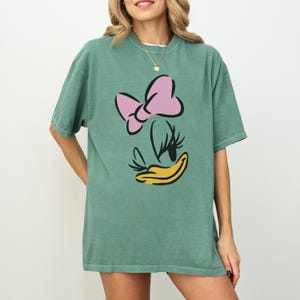 May include: A green t-shirt featuring a cartoon image of Daisy Duck. The design includes a pink bow, eyelashes, and a yellow beak. The shirt has short sleeves and a relaxed fit.
