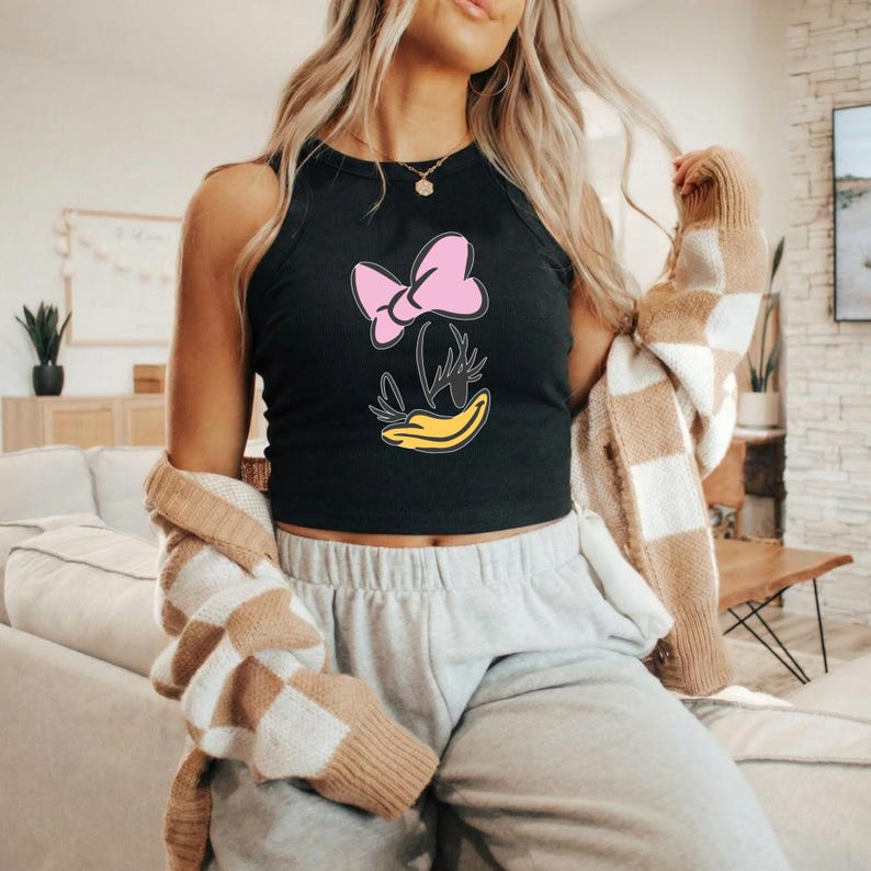 Op de afbeelding: Zwarte tanktop met een roze strik en een gele eendenbek-ontwerp. De mouwloze top heeft een cartoonillustratie. De top wordt gedragen met grijze joggingbroek en een bruin-wit geruit vest.