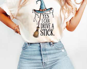 Puede incluir: Camiseta color crema con un diseño de sombrero de bruja y escoba con el texto "YES I CAN DRIVE A STICK." El sombrero es azul con detalles marrones, y la escoba es marrón. La camiseta se combina con vaqueros azules claros.