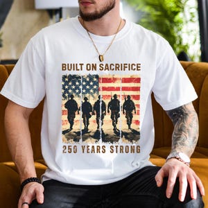 May include: White t-shirt featuring an American flag design with soldiers and the text "BUILT ON SACRIFICE" and "250 YEARS STRONG". The design incorporates red, white, and blue colors, with a vintage, distressed aesthetic.