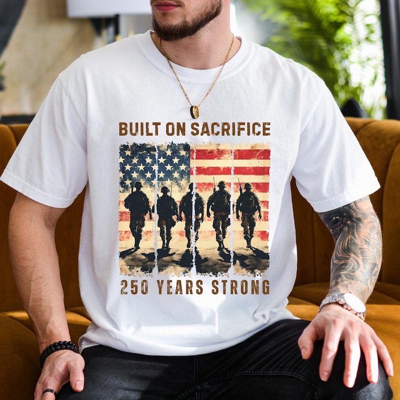 May include: White t-shirt featuring an American flag design with soldiers and the text "BUILT ON SACRIFICE" and "250 YEARS STRONG". The design incorporates red, white, and blue colors, with a vintage, distressed aesthetic.