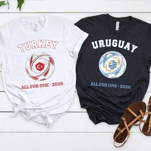 May include: Two t-shirts with soccer ball graphics. One white shirt has "TURKEY" and a soccer ball with a Turkish flag design, and the text "ALL FOR ONE 2026". The other is a dark gray shirt with "URUGUAY" and a soccer ball with an Uruguayan flag design, and the text "ALL FOR ONE 2026".