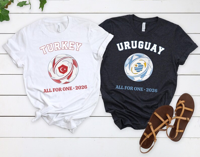 May include: Two t-shirts with soccer ball graphics. One white shirt has "TURKEY" and a soccer ball with a Turkish flag design, and the text "ALL FOR ONE 2026". The other is a dark gray shirt with "URUGUAY" and a soccer ball with an Uruguayan flag design, and the text "ALL FOR ONE 2026".