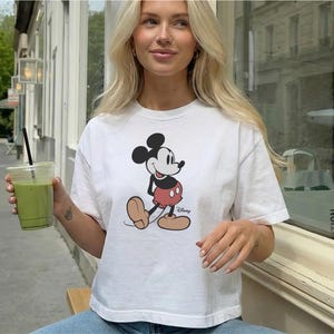 以下が含まれることがあります： ミッキーマウスのデザインが施された白いTシャツ。Tシャツは丸首で半袖です。画像には、黒いストローが入った透明なプラスチックカップに入った緑色の飲み物も写っています。