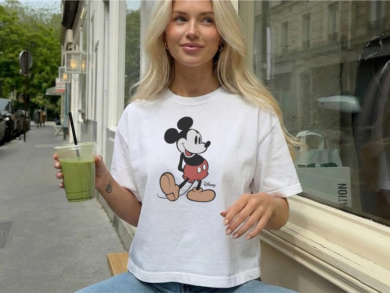 以下が含まれることがあります： ミッキーマウスのデザインが施された白いTシャツ。Tシャツは丸首で半袖です。画像には、黒いストローが入った透明なプラスチックカップに入った緑色の飲み物も写っています。