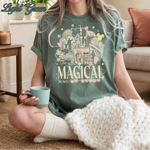 May include: A light green t-shirt featuring a graphic print with various park-themed icons, including a castle, a monorail, and a teacup. The text reads "The Most MAGICAL PLACE" in a stylized font, surrounded by stars, flowers, and heart shapes.
