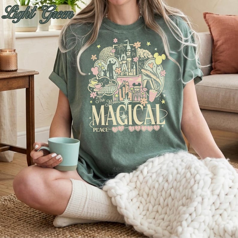 May include: A light green t-shirt featuring a graphic print with various park-themed icons, including a castle, a monorail, and a teacup. The text reads "The Most MAGICAL PLACE" in a stylized font, surrounded by stars, flowers, and heart shapes.