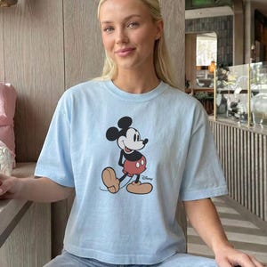 以下が含まれることがあります： ミッキーマウスのグラフィックが特徴のライトブルーのTシャツ。漫画のキャラクターは、赤いショートパンツ、黄色の靴、白い手袋を着用した、彼のクラシックなポーズで描かれています。カジュアルなスタイルです。
