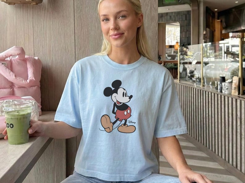 以下が含まれることがあります： ミッキーマウスのグラフィックが特徴のライトブルーのTシャツ。漫画のキャラクターは、赤いショートパンツ、黄色の靴、白い手袋を着用した、彼のクラシックなポーズで描かれています。カジュアルなスタイルです。