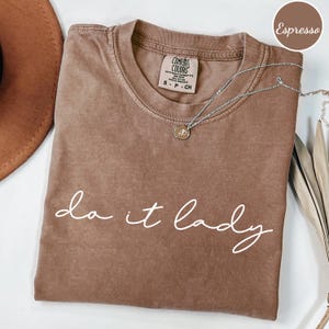 Puede incluir: Camiseta color espresso con la frase "do it lady" en escritura blanca. La camiseta tiene cuello redondo y mangas cortas. Un collar plateado con un peque&ntilde;o colgante est&aacute; drapeado sobre la camiseta.