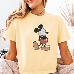 以下が含まれることがあります： ミッキーマウスのイラストが描かれた、明るい黄色のTシャツ。漫画のキャラクターは、赤いショートパンツ、白い手袋、茶色の靴というクラシックな衣装で描かれています。ディズニーのロゴが見えます。